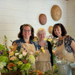Three women wearing beige aprons smile behind a table filled with a large basket of fresh flowers in a bright, rustic indoor setting with white shiplap walls and woven wall decor.
