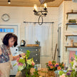 A woman in a floral studio arranging flowers on a rustic wooden table, surrounded by colorful blooms, with a white shiplap wall and wooden ceiling in the background.
