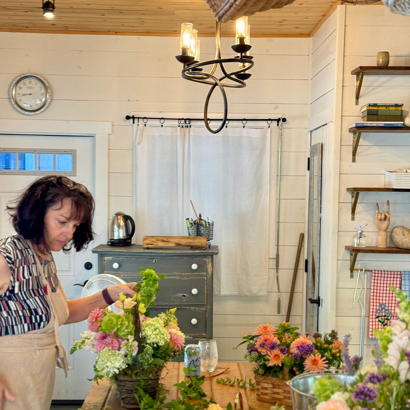 A woman in a floral studio arranging flowers on a rustic wooden table, surrounded by colorful blooms, with a white shiplap wall and wooden ceiling in the background.