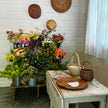 A rustic styled interior scene with a white shiplap wall, decorative woven wall plates, a floral arrangement display in metal buckets, a distressed white table with a round wooden board, and two woven baskets near a window with sheer white curtains.