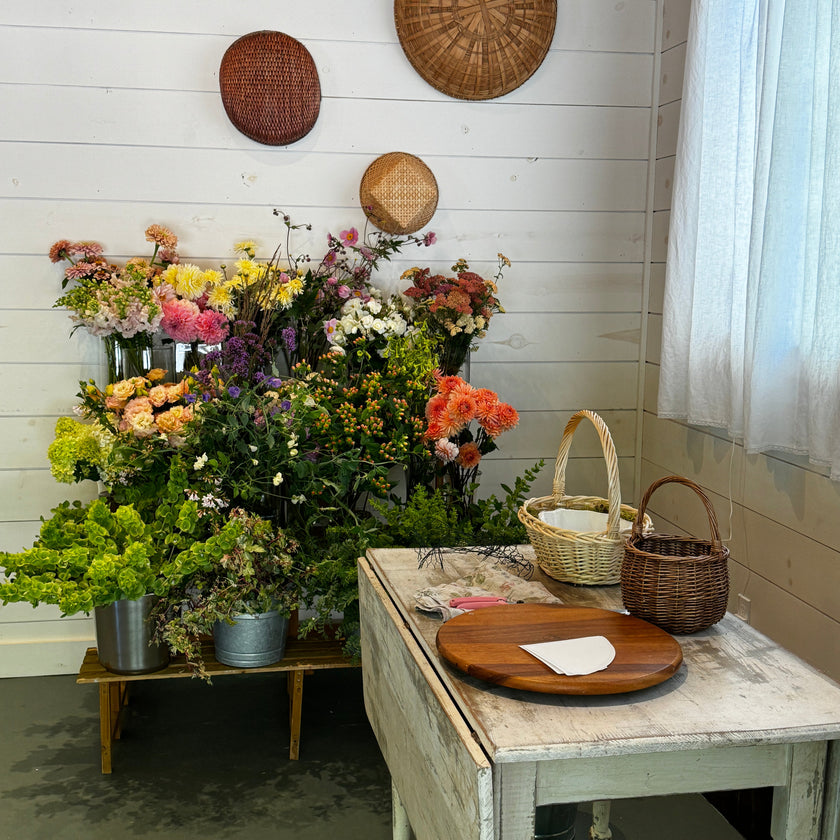 A rustic styled interior scene with a white shiplap wall, decorative woven wall plates, a floral arrangement display in metal buckets, a distressed white table with a round wooden board, and two woven baskets near a window with sheer white curtains.