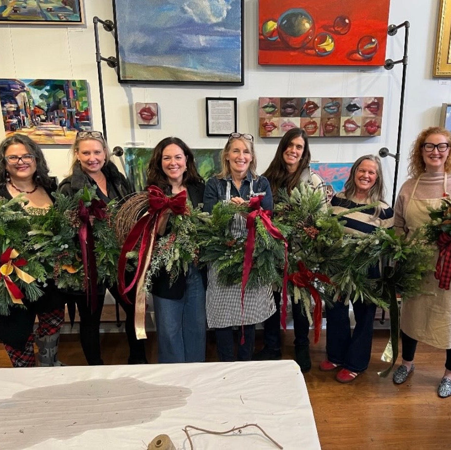 A group of seven women standing indoors and holding handmade green wreaths with red ribbons and bows. The room has framed artwork on the walls, a wooden floor, and a table in the foreground.