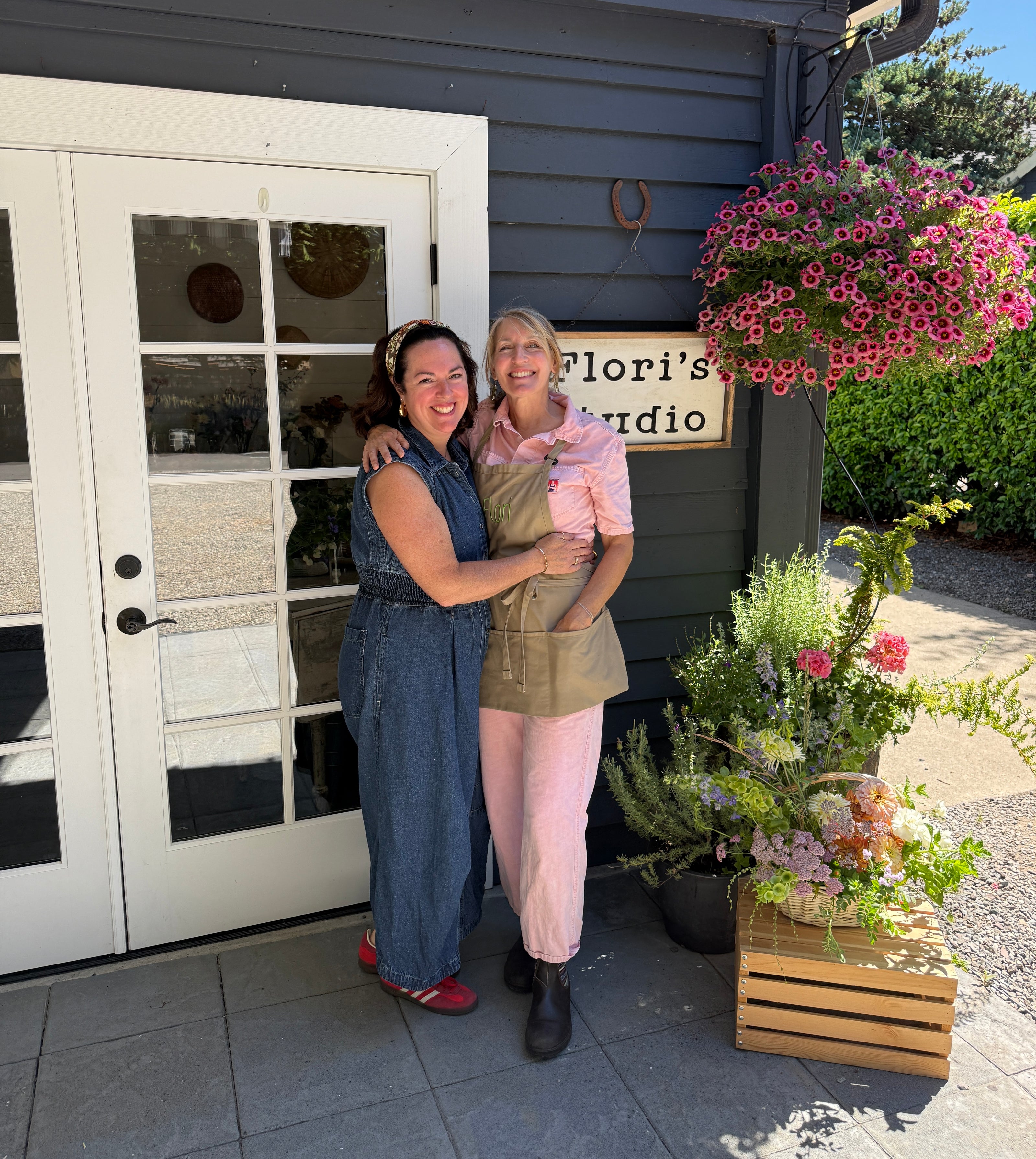 Two women posing together outdoors in front of a building with dark gray siding and white double glass doors. One wears a blue denim jumpsuit, and the other wears a pink top, tan apron, and pink pants.