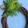 A rustic circular grapevine wreath made of intertwined brown branches, decorated with green evergreen foliage and a spool of twine, set against a light blue background.
