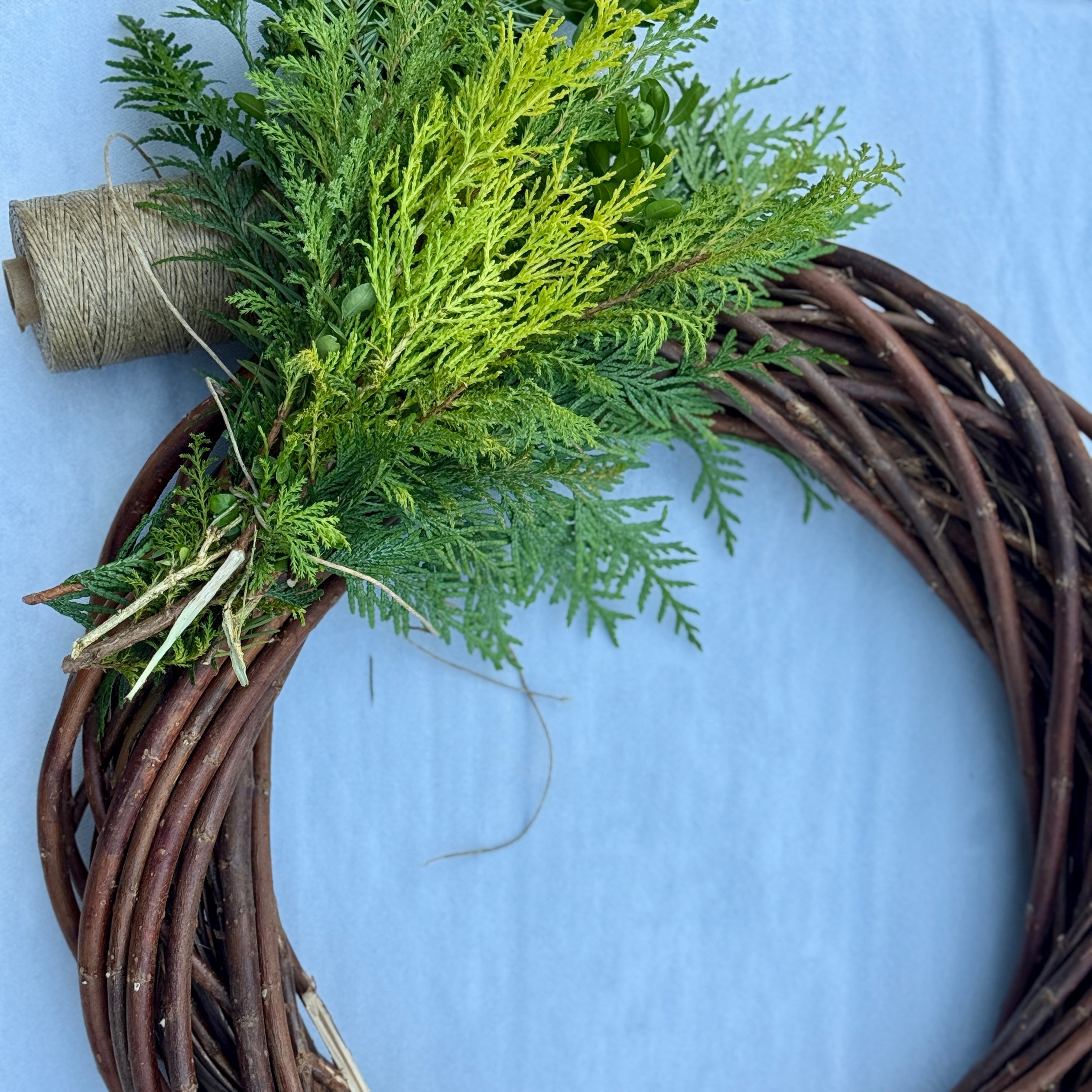 A rustic circular grapevine wreath made of intertwined brown branches, decorated with green evergreen foliage and a spool of twine, set against a light blue background.